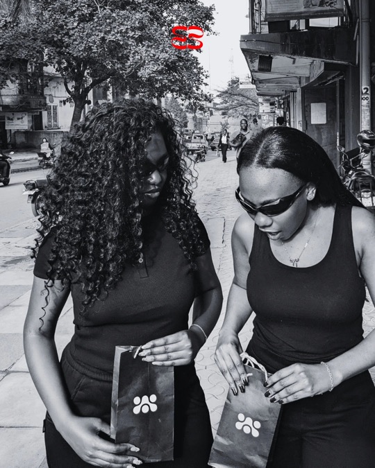 Two women in black looking into ruvori paper bags on the pavement, in black and white.