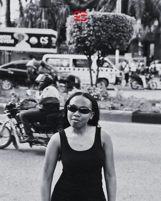 A smiling woman in ruvori sunglasses, a motorbike rider blurred behind her, in black and white.