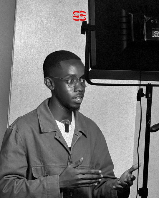 Behind the scenes: a man in a brown jacket and round glasses, a small clip microphone on his collar, speaks beside a studio light, in black and white.