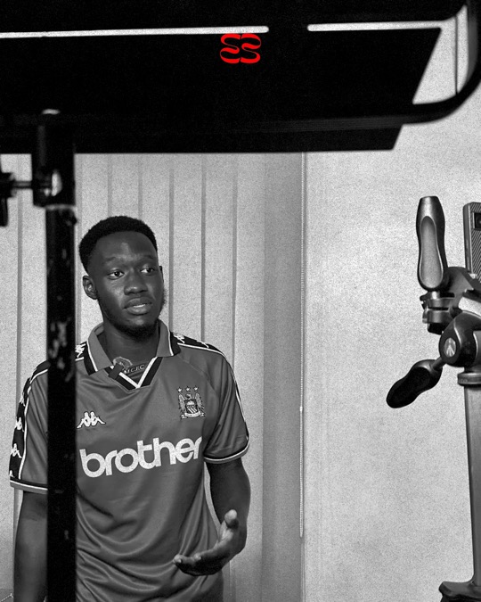 Behind the scenes: a man in a football shirt with a clip microphone talks on set between a studio light stand and a camera tripod, in black and white.