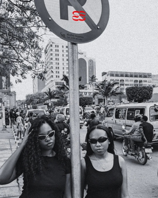 Two women in ruvori sunglasses under a no-parking sign on a busy Kampala street, in black and white.