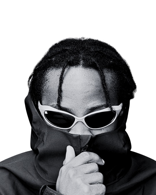 Black and white portrait of a man with braids, a dark jacket zipped up to his nose, wearing white-framed Ruvori sunglasses.