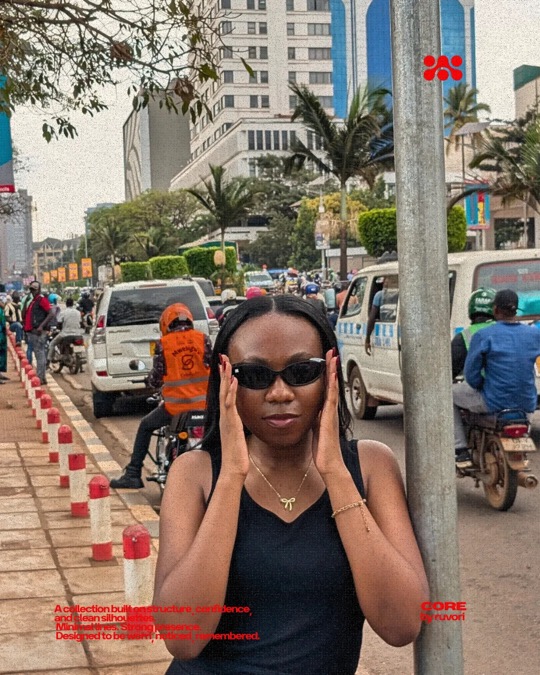 Woman in black Ruvori sunglasses and a black top, hands held to her face, leaning on a pole beside a busy road with motorbike taxis and a white minibus.